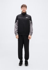 Μπουφάν Puma για τρέξιμο σε μαύρο και γκρι, με φερμουάρ μπροστά και ελαστικά μανίκια, σε συνδυασμό με μαύρο παντελόνι. Το υλικό φαίνεται λείο με εφαρμοστή σχεδίαση.