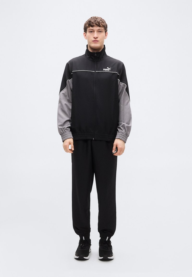 Μπουφάν Puma για τρέξιμο σε μαύρο και γκρι, με φερμουάρ μπροστά και ελαστικά μανίκια, σε συνδυασμό με μαύρο παντελόνι. Το υλικό φαίνεται λείο με εφαρμοστή σχεδίαση.