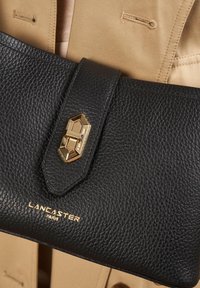 Clutch in pelle nera con superficie testurizzata, chiusura in metallo dorato e logo "LANCASTER PARIS" in oro. Dettaglio della chiusura a forma triangolare.