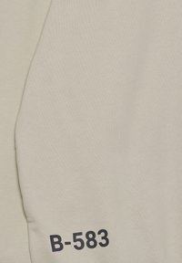 Felpa beige chiaro con una texture liscia, caratterizzata da una stampa audace "B-583" di colore blu navy su una manica. Design semplice, stile informale.