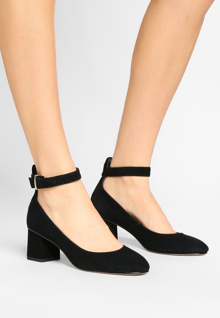 Chaussures à talon bloc en daim noir avec bride cheville portées pieds nus sur un fond blanc uni.