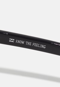 Schwarze Sonnenbrille mit glänzender Oberfläche, mit dem aufgedruckten weißen Text "KNOW THE FEELING" und einem kleinen Logo am Bügel.