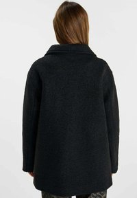 Zwarte wollen jas met een brede kraag, dikke textuur en een losse pasvorm, met zichtbare naden en een middellange snit.