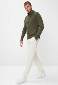 Mann, der geht und einen olivgrünen Reißverschluss-Pullover, hellbeige Hose und weiße Sneakers trägt, vor einem schlichten hellen Hintergrund.