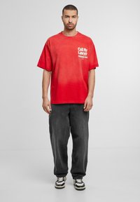 Rød T-shirt med gradienteffekt og hvid tekst på forsiden, kombineret med løstsiddende sorte bukser og sort-hvide sneakers.