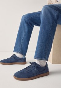 Blå ruskindssneakers med en gummisål, der har en lav profil og snørelukning, kombineret med op rullede denimjeans og hvide sokker.