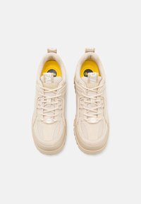 Zapatillas deportivas beige con una parte superior de ante texturizado y malla, con una plantilla amarilla y un logo destacado en la lengüeta y el talón.