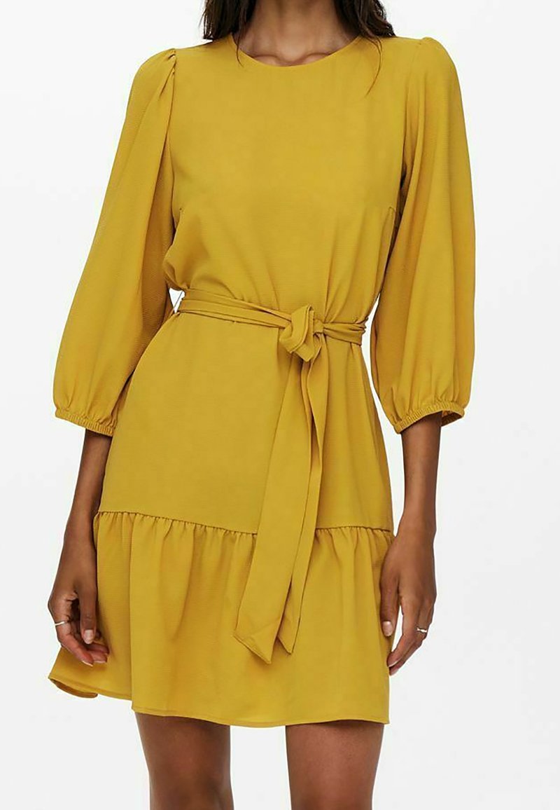 Femme portant une robe jaune moutarde longueur genou avec manches bouffantes trois-quarts et ceinture nouée à la taille.