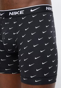 Μαύρα boxer briefs με πολλαπλά λευκά λογότυπα της Nike σε σχήμα φτερού και μαύρη μέση που φέρει το όνομα Nike και το λογότυπο φτερού σε λευκό.