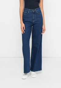 Jeans de ganga azul de perna larga com cintura alta, cinco bolsos e bainha limpa. Combinados com ténis brancos.