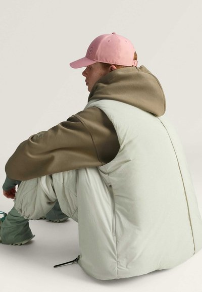 Helles Mint-farbenes Puffer-Weste mit einem braunen Kapuzenpullover darunter, kombiniert mit lockeren, strukturierten Hosen und einer pinken Mütze mit einem Logo.