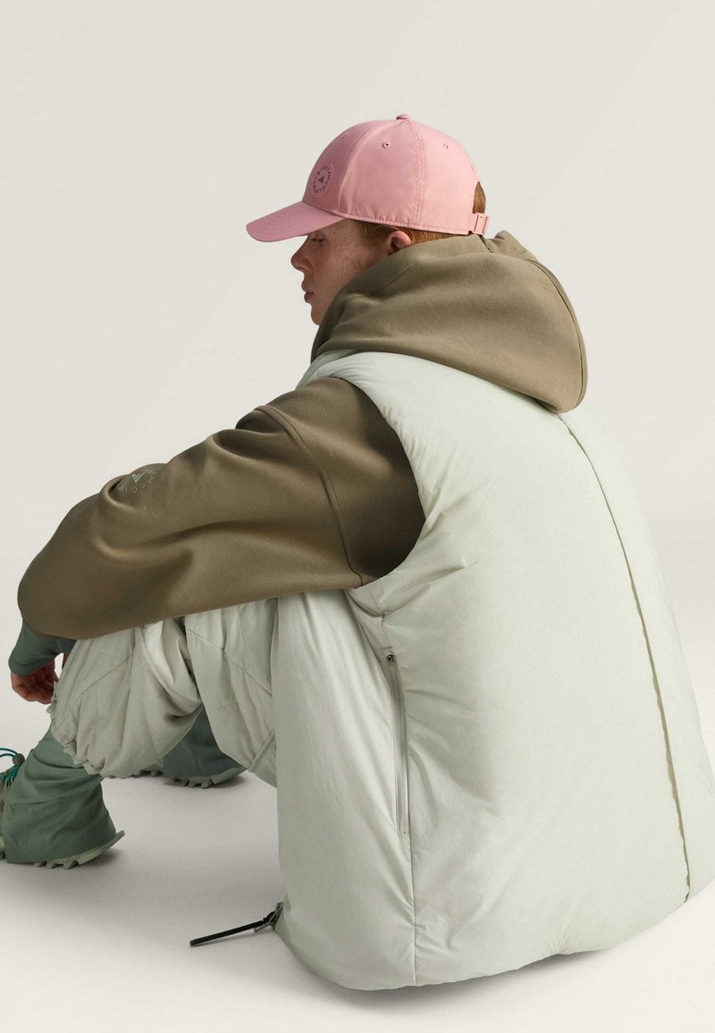 Chaleco de pluma mint claro con una sudadera con capucha marrón debajo, combinado con pantalones sueltos y texturizados y una gorra rosa con un logotipo.