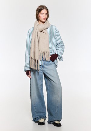 Bežna volnena ruta z resicami, oversize črtasta srajca v beli in zeleni barvi ter široke modre jeans hlače, dopolnjene s črnimi čevlji.