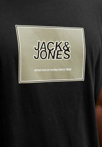 Sort bomuld t-shirt med en stor, rektangulær grafik med teksten "JACK & JONES" i fed sort skrift og en tekstureret grøn baggrund.