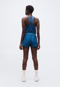 Άτομο που φαίνεται από πίσω, φοράει μπλε αθλητική φανέλα και σορτς, λευκές κάλτσες και λευκά αθλητικά παπούτσια, με φόντο απλό και φωτεινό.