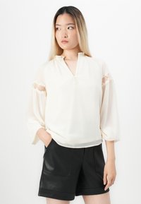 Kvinna iklädd en krämfärgad blus med spetskant på ärmarna och svarta lädershorts, stående med en hand i fickan, tittande åt vänster.