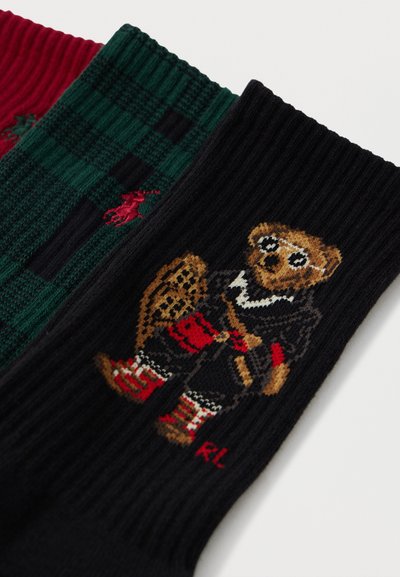 Trīs zeķu pāri: sarkanas ribotas, zaļas rūtainas un melns ar rotaļlietu lāci sporta tērpā. Materiāls šķiet adīts, ar krāsainām detaļām.