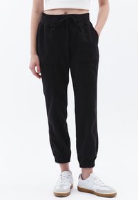 Zwarte, lichte, taps toelopende broek met een elastische tailleband, trekkoord en twee grote voorkruiszakken. De stof lijkt zacht met een gladde textuur.