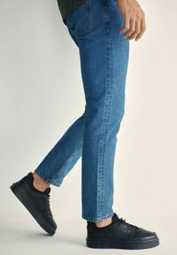 Osoba w niebieskich dżinsach i czarnych trampkach z sznurowanym designem, stojąca na jednolitym jasnym tle.