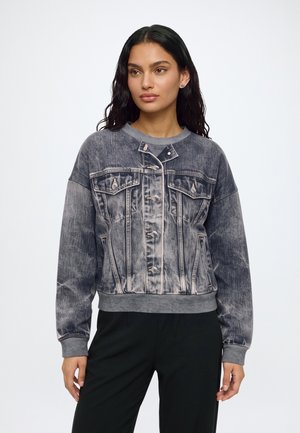 Femme aux longs cheveux foncés portant une veste en jean noire délavée boutonnée et un pantalon noir sur un fond blanc uni.