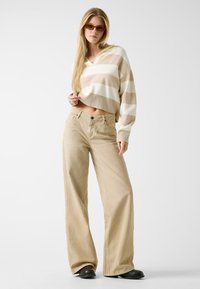 Pull court rayé beige et blanc avec un col en V, associé à un pantalon kaki à jambes larges et des chaussures noires.