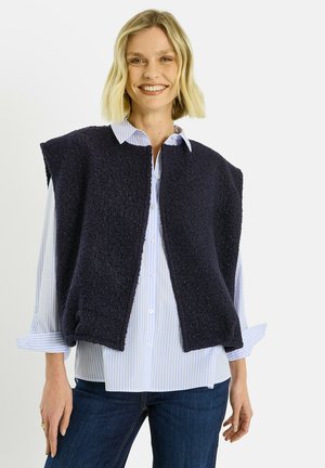 Chaleco de forro polar azul marino con cuello redondeado y sin mangas, combinado con una camisa de botones de rayas azul claro, y pantalones vaqueros oscuros.