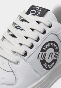 Λευκό sneaker Versace Couture Jeans με λευκά κορδόνια, μαύρο λογότυπο στο πλάι και μαύρη ετικέτα λογότυπου στη γλώσσα.