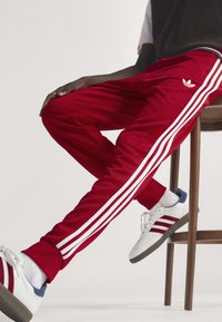 Άτομο που φοράει κόκκινη φόρμα Adidas με λευκές ρίγες και λευκά αθλητικά παπούτσια, κάθονται σε ένα ξύλινο σκαμπό μπροστά σε απλό φόντο.