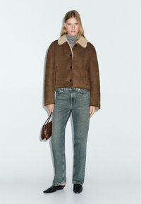 Bruine suède croptop jas met een beige bontkraag, knoopsluitingen, gecombineerd met vervaagde blauwe jeans en zwarte instap schoenen.