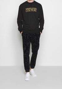 Svart sweatshirt med guld "Versace Jeans Couture" logotyp, kombinerad med svarta joggers med blommönster och vita sneakers. Mjuk tygkvalitet, avslappnad passform.