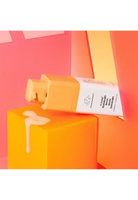 A tube of serum with an orange cap, white body, and text, resting on a bright yellow cube with a drip of serum on the surface.