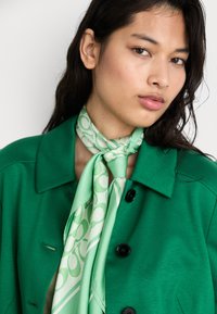 Green jacket with black buttons, paired with a light green patterned scarf tied at the neck featuring abstract shapes and stripes.