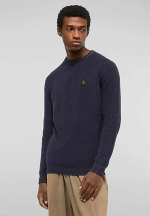 Uomo che indossa un maglione a coste blu navy a maniche lunghe con una piccola toppa sul petto, pantaloni beige, in piedi contro uno sfondo grigio chiaro uniforme.