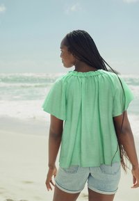 Blusa leggera di colore verde menta con scollatura arricciata e maniche larghe, abbinata a pantaloni shorts in denim chiaro. Indossata da una persona che guarda verso il mare.