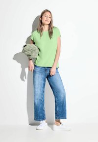 Top verde a maniche corte, jeans larghi blu e sneakers bianche. La modella tiene una borsa verde intrecciata e testurizzata con manici.