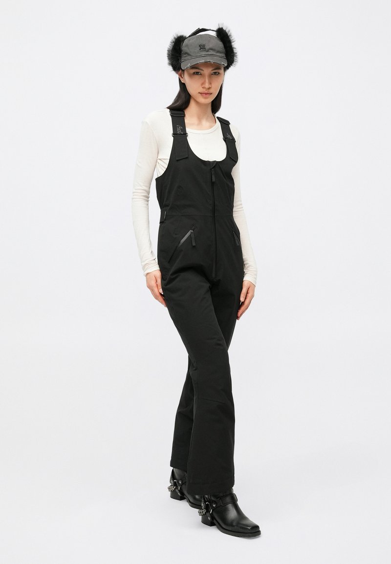 Black ski overalls with adjustable straps, front zipper, and two zippered pockets. Worn over a white long-sleeve top and paired with a gray fur-accented cap.