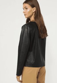 Mujer con cabello largo castaño, vistiendo una chaqueta de cuero negra y pantalones beige, mirando hacia la izquierda contra un fondo claro y liso.