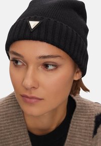Cappello a maglia nero con design a costine e una toppa a forma di triangolo sul davanti. Presenta una vestibilità aderente e una texture morbida.