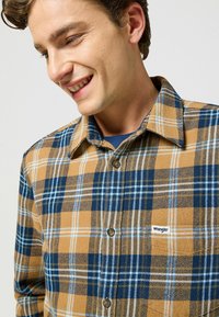 Brun och blå rutigt skjorta tillverkad av flanelltyg. Har en knapp-up krage och en liten logotypetikett. Texturerat mönster med kontrasterande färger.