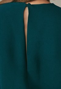 Ingrandimento della scollatura posteriore di un indumento teal con una piccola apertura a chiave fissata da un bottone rivestito di stoffa.