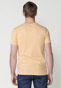 Krátký rukáv, tričko s kulatým výstřihem z lehkého oranžového materiálu s jemnou texturou. Pohled zezadu; žádné viditelné značení ani vzory. Nosí se s tmavými džíny.