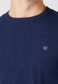 Blå stickad tröja med rund halsringning och en liten, ljusblå logotyp på bröstet. Mjuk textur och jämn sömnad genom hela tröjan.