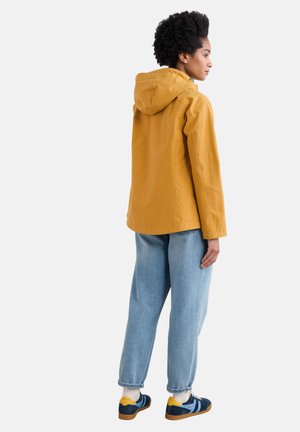 Veste à capuche jaune avec une coupe décontractée, associée à un jean en denim bleu clair et des baskets bleues avec des accents jaunes. Style doux et décontracté.
