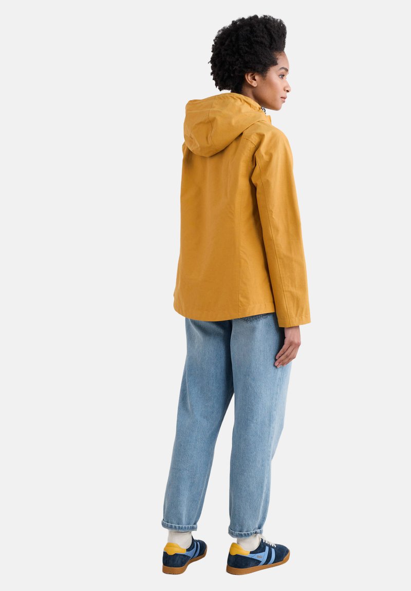 Gele capuchonjack met een losse pasvorm, gecombineerd met lichtblauwe denim jeans en blauwe sneakers met gele accenten. Zachte, casual stijl.