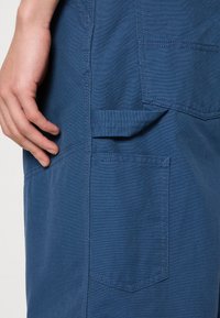 Vasen käsi lepää sinisten ty�öhousujen vieressä, joissa on useita ommeltuja taskuja ja kankainen lenkki sivussa.