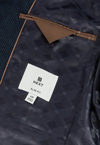 Doublure intérieure d’une veste slim fit à motif foncé montrant une poche marron et une étiquette blanche avec la marque NEXT et la taille 40R.