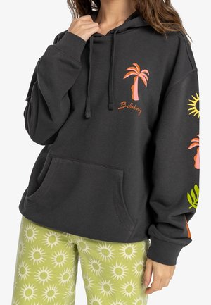 Osoba w czarnej bluzie z kapturem z pomarańczowym obrazem palmy i zielonych spodniach z białymi wzorami słońca, stojąca na białym tle.