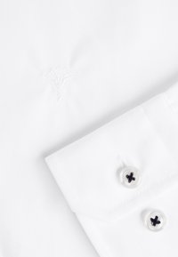 Camisa blanca de vestir con un logo bordado, presenta un cuello puntiagudo y puño abotonado con botones oscuros contrastantes y textura suave.