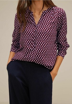Žena s fialovo-růžovou geometricky vzorovanou košilí s vyhrnutými rukávy, v kombinaci s černými kalhotami a rukama v kapsách.