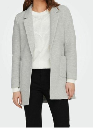 Femme portant un blazer gris clair sur un pull en maille blanc et un pantalon noir, montrée du cou jusqu'à mi-cuisse sur un fond uni.